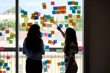 Two people discussing coloured post-it notes stuck to a window with the sun shining in